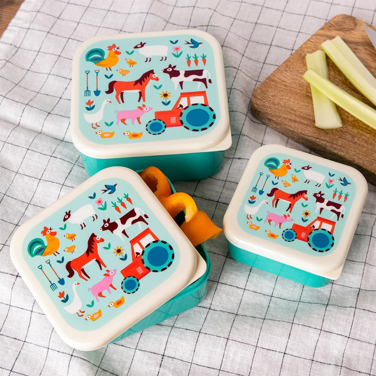 Three colorful children's lunch boxes with farm animal designs on a checkered tablecloth.