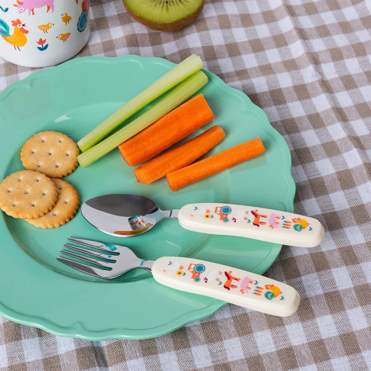 Children's plate with crackers, carrots, celery, and floral-patterned fork and spoon on a checkered tablecloth.