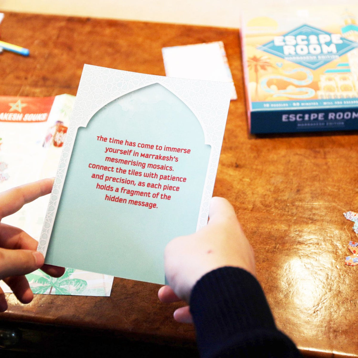 Person holding a card with text on a wooden table with an 'Escape Room' game box in the background.