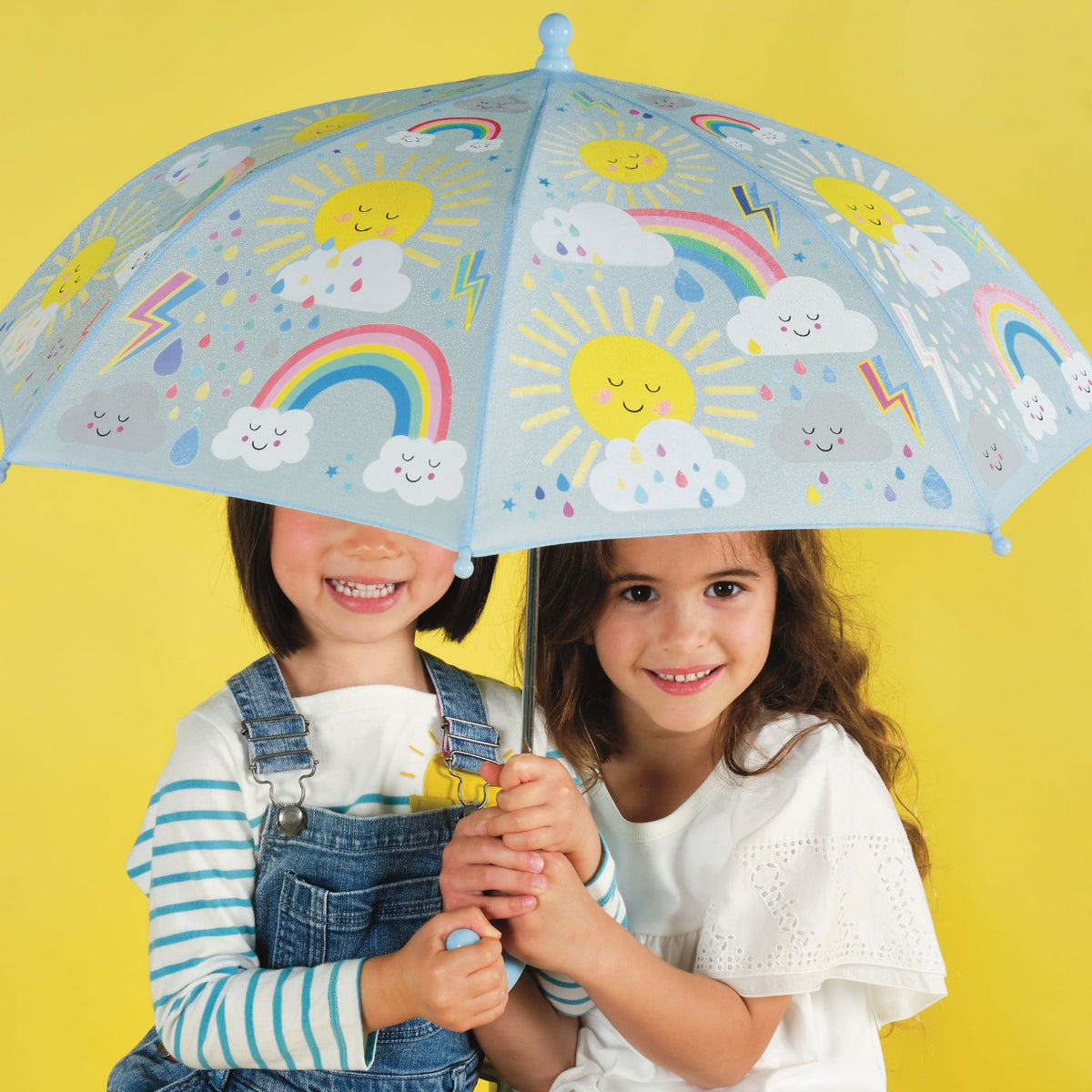 Two children holding a colorful umbrella with sun, rainbow, and cloud designs
