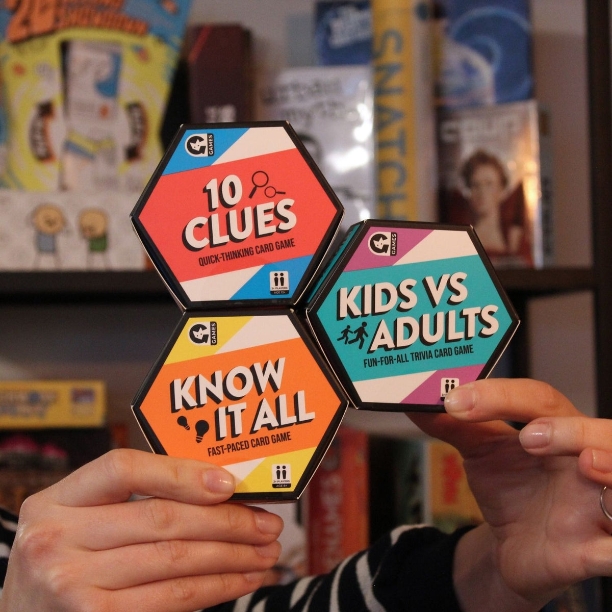Three card game boxes titled '10 Clues', 'Kids vs Adults', and 'Know It All' held by a person against a blurred background of more card games.