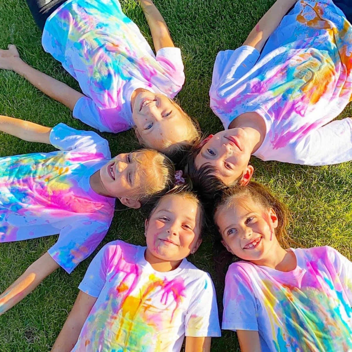five kids lying on the grass looking up at the camera in a circle. They are wearing white tshirts and covered in paint. 