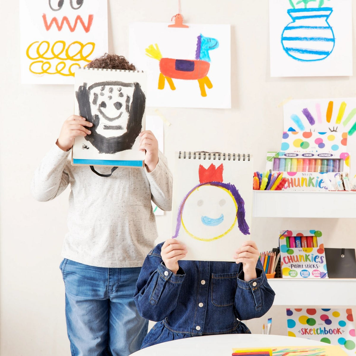 Two children holding up their homemade masks and colourful drawings made with their chunkies paint sticks