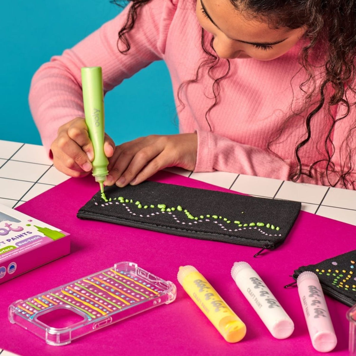Child using a green dot-a-lot paint marker to draw on a black pouch with colorful markers and cases on a pink surface.