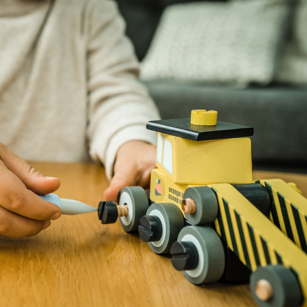 A close-up shot of a young child’s hands carefully connecting and screwing the wooden body pieces of the digger on a low play table, capturing concentration and fine motor focus.